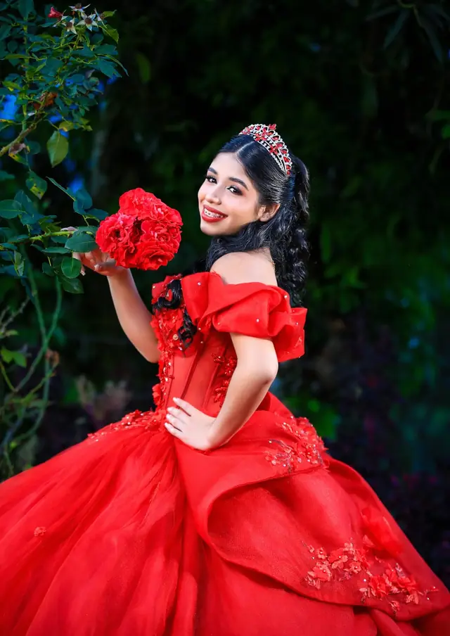 Quinceañera con vestido rojo que sostiene un ramo de flores rojas en un jardín